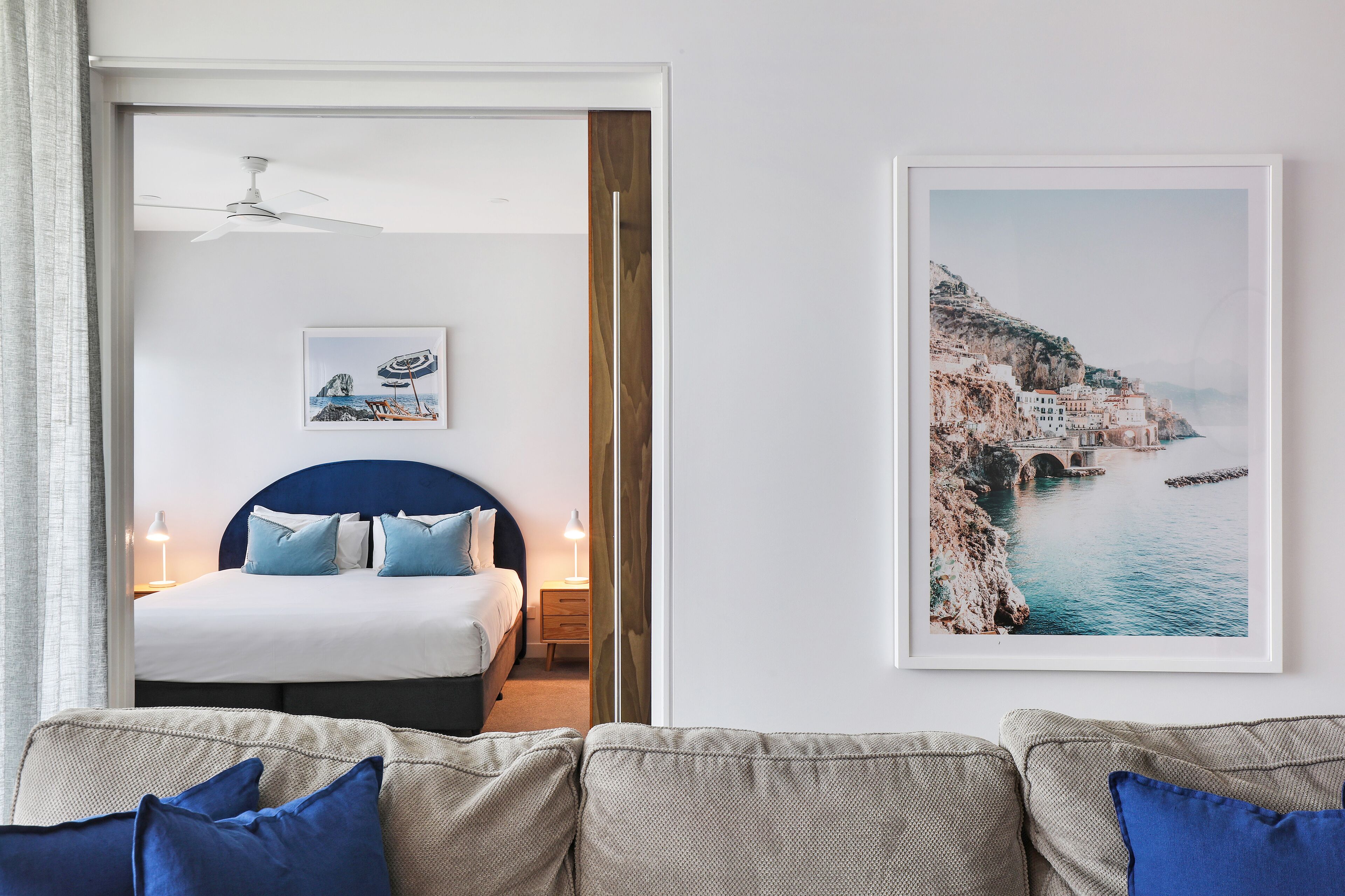 Un dormitorio con una cama, mesitas de noche, un ventilador de techo y un cuadro enmarcado con un paisaje costero en la pared.
