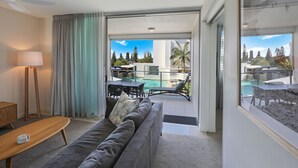 Un salon moderne avec un canapé gris, une table basse en bois et un lampadaire. Une porte-fenêtre coulissante donne accès à un espace extérieur doté d’une piscine et de chaises longues.