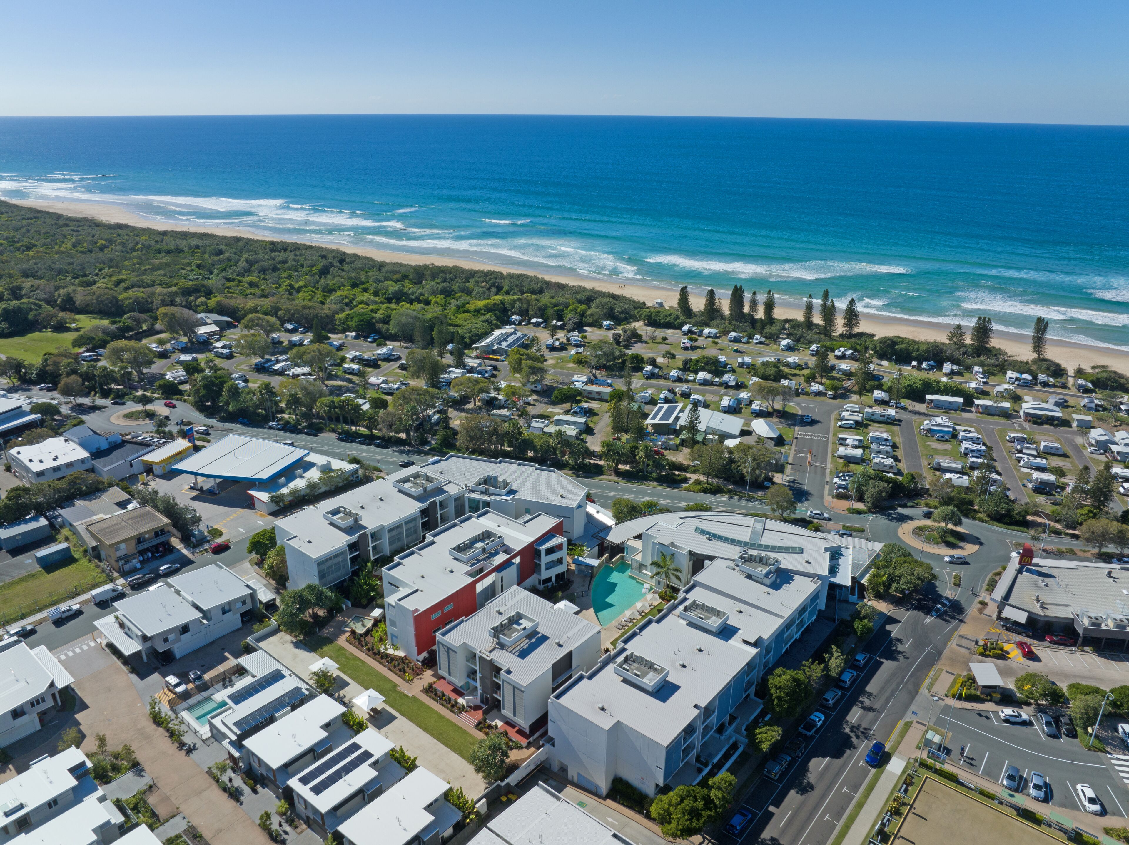 An aerial view of a coastal residential area with modern buildings, a beach, and a clear blue sky.
