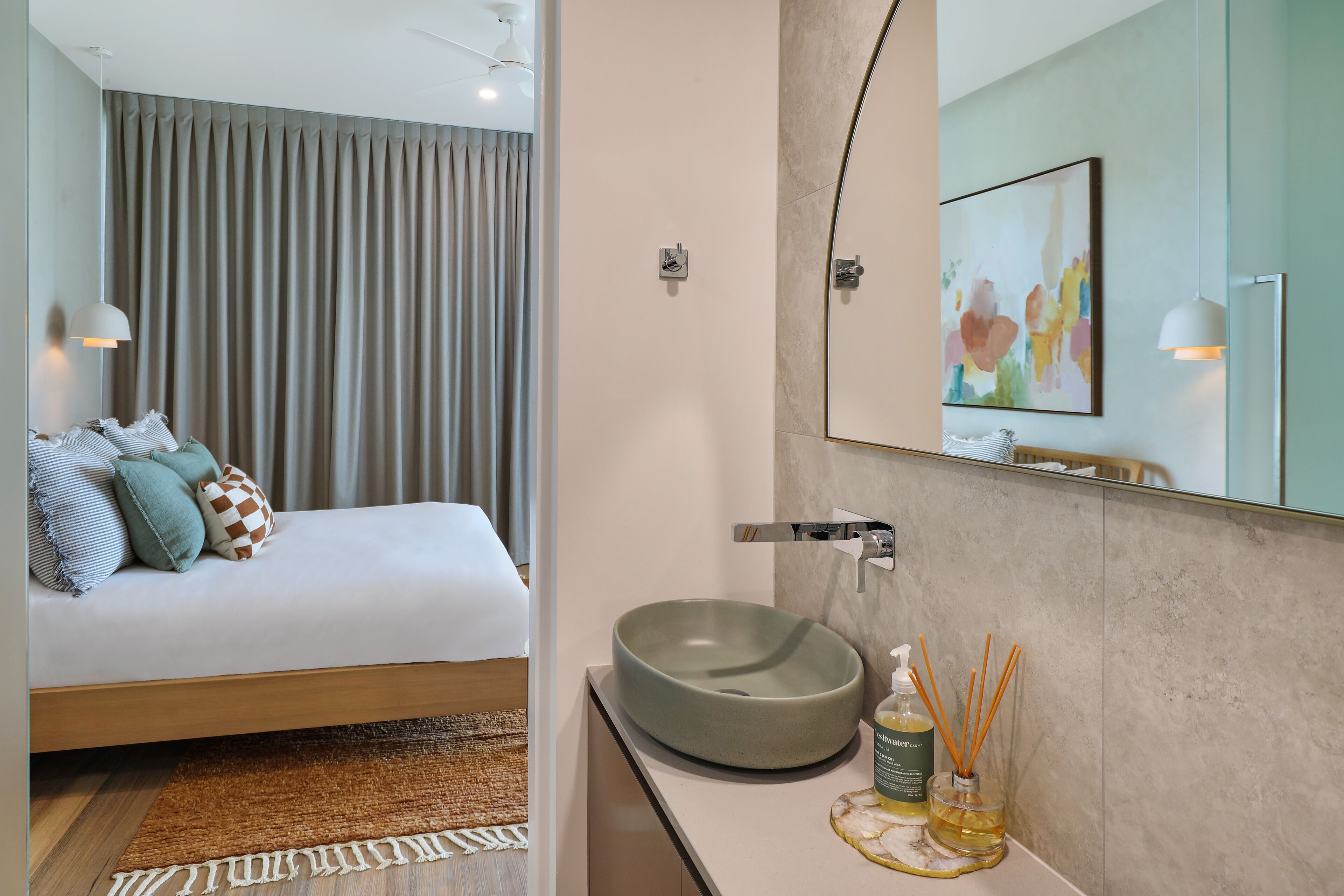 Une chambre à coucher moderne avec un lit, des oreillers et une lampe de chevet. La salle de bain est équipée d’un lavabo, d’un miroir et d’une douche.