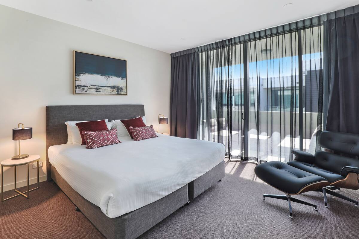 A modern hotel room with a large bed, a side table with a lamp, a black leather chair, and a view of the city through sheer curtains.