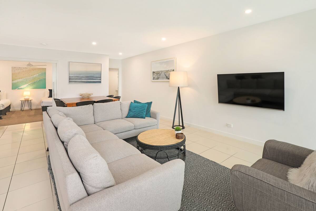 A modern living room with a grey sectional sofa, a round coffee table, and a flat-screen TV mounted on the wall.