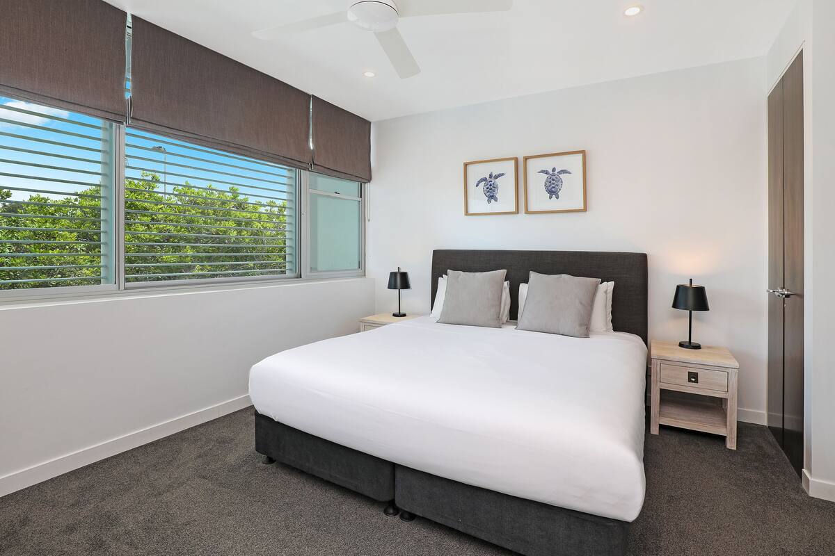 A modern bedroom with a large bed, two bedside tables, a ceiling fan, and a window with blinds.