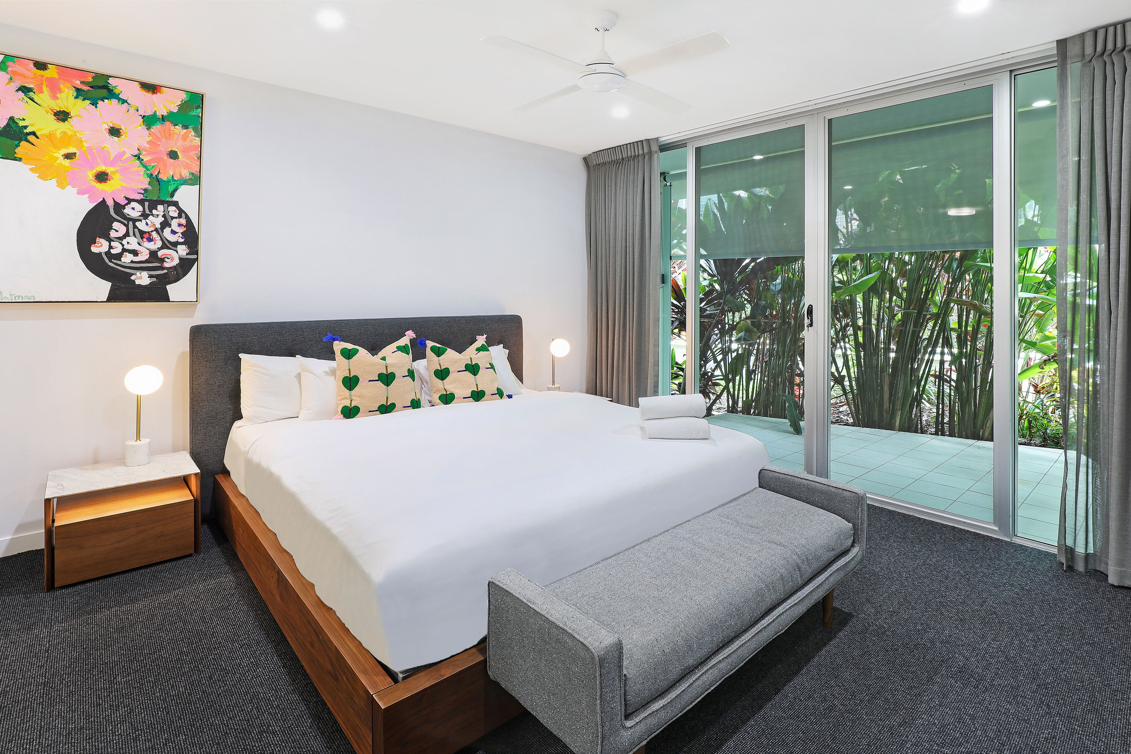 Una habitación de hotel moderna con una cama grande, una banca, una mesita de noche con lámpara, un ventilador de techo y un ventanal con vistas a zonas verdes.