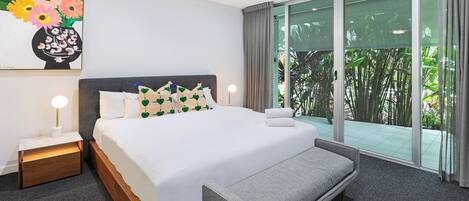 Una habitación de hotel moderna con una cama grande, una banca, una mesita de noche con lámpara, un ventilador de techo y un ventanal con vistas a zonas verdes.