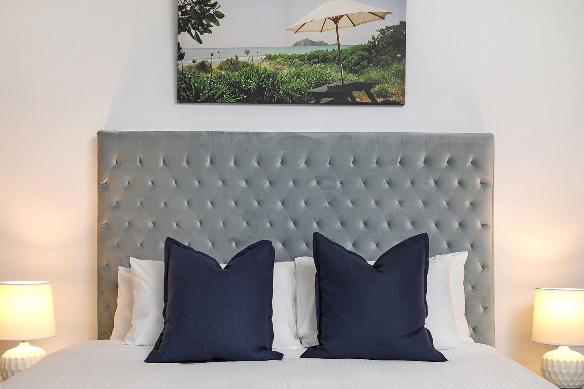 A bedroom with a tufted headboard, a bed with white and navy pillows, and a framed landscape painting above the bed.