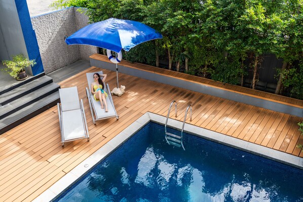 Una terraza de madera con piscina, sillas de descanso y una sombrilla azul.