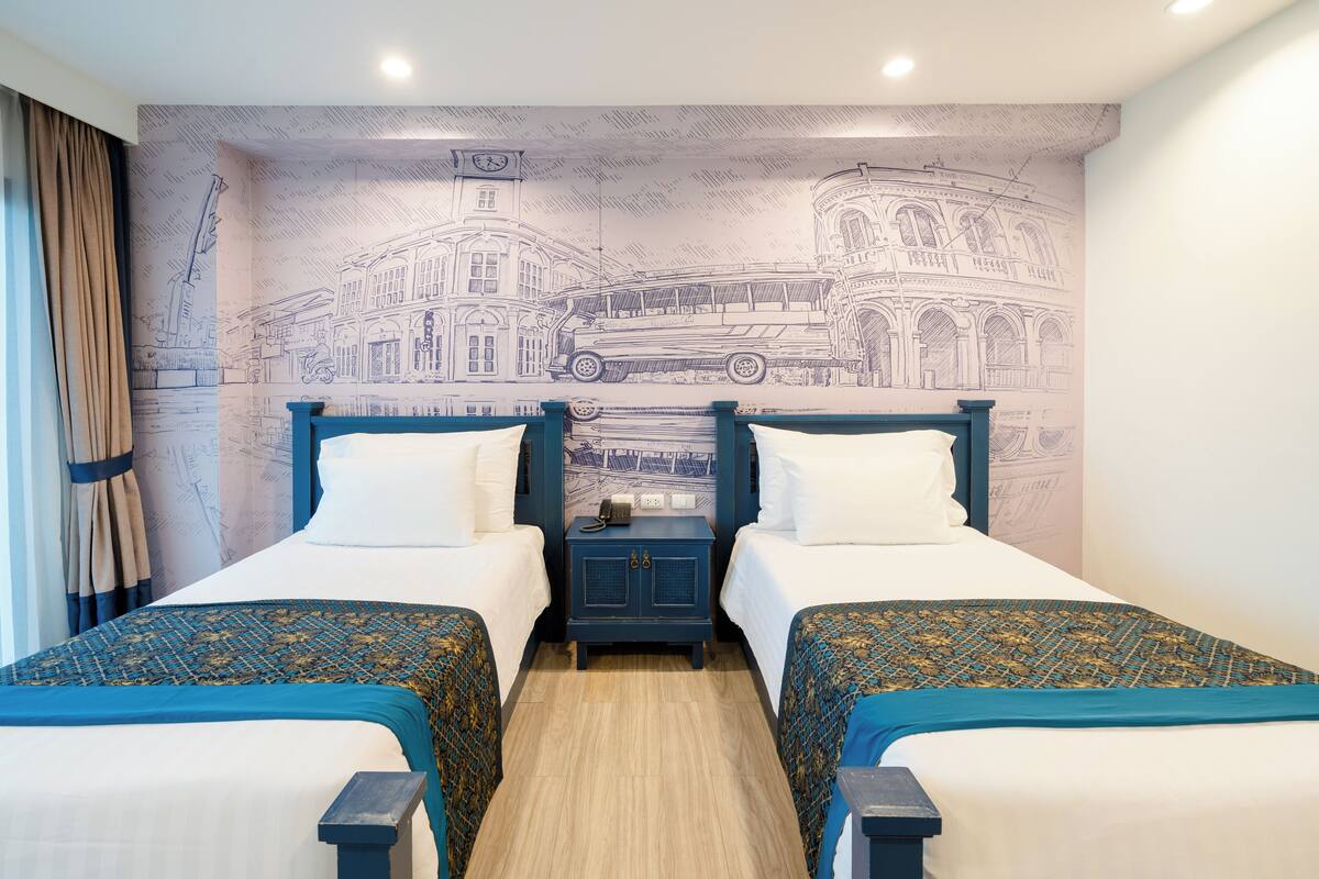 Habitación de hotel con dos camas, una mesita de noche y un mural con un paisaje urbano en la pared.