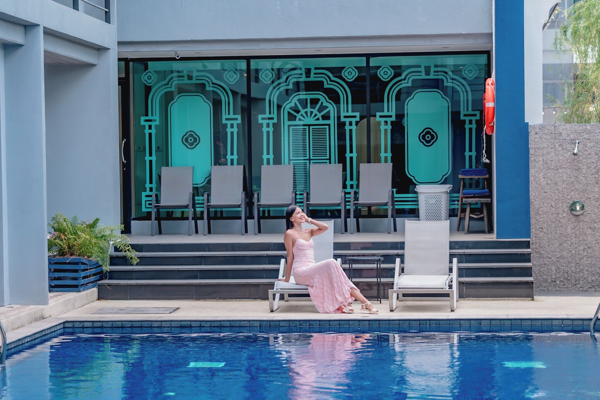 Una mujer con un vestido rosa sentada junto a una piscina, con sillas de descanso y un edificio con amplias ventanas al fondo.