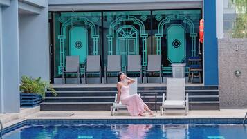 Una mujer con un vestido rosa sentada junto a una piscina, con sillas de descanso y un edificio con amplias ventanas al fondo.