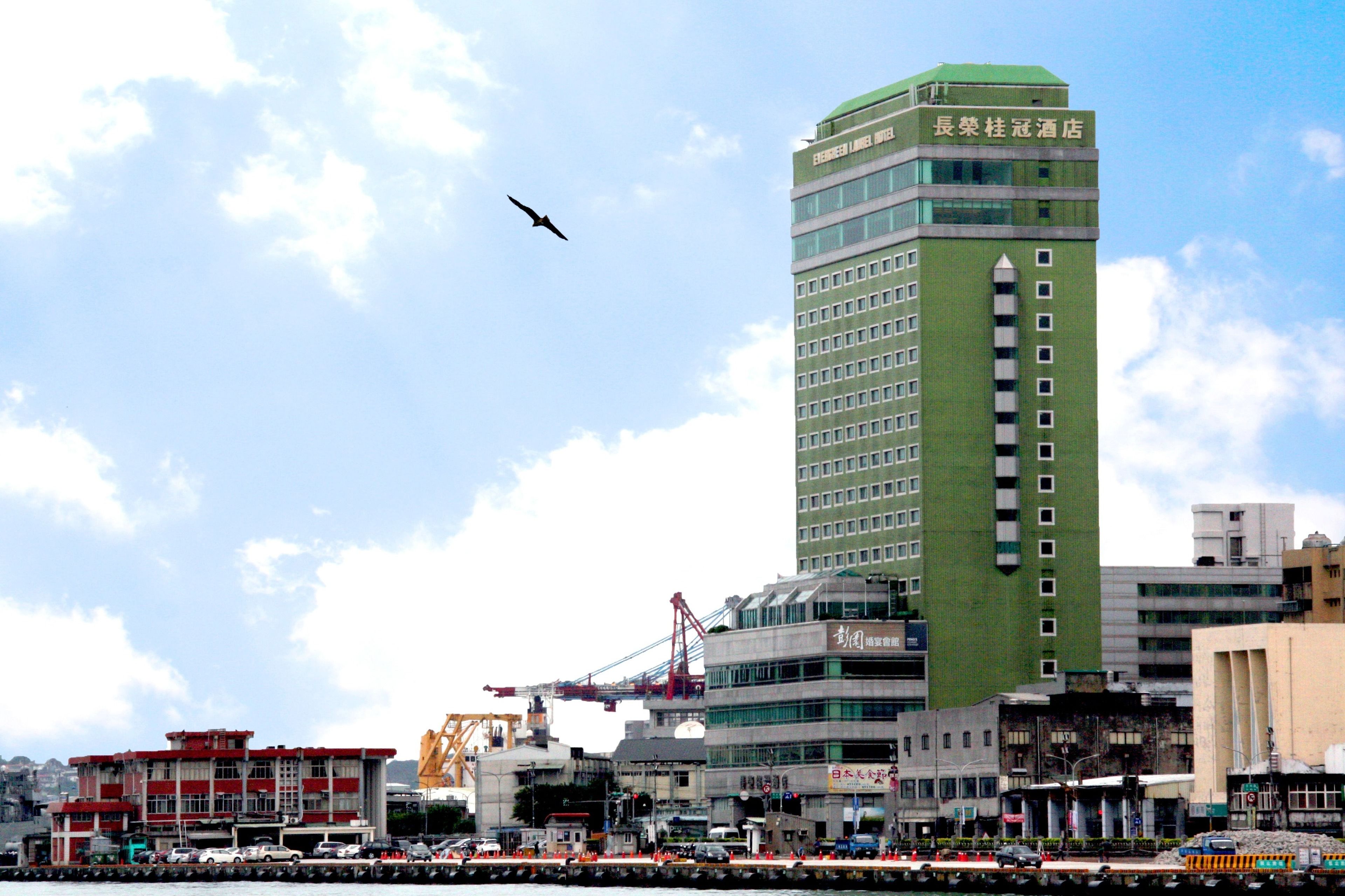 A tall hotel with multiple floors and a green facade, surrounded by other buildings and construction cranes.