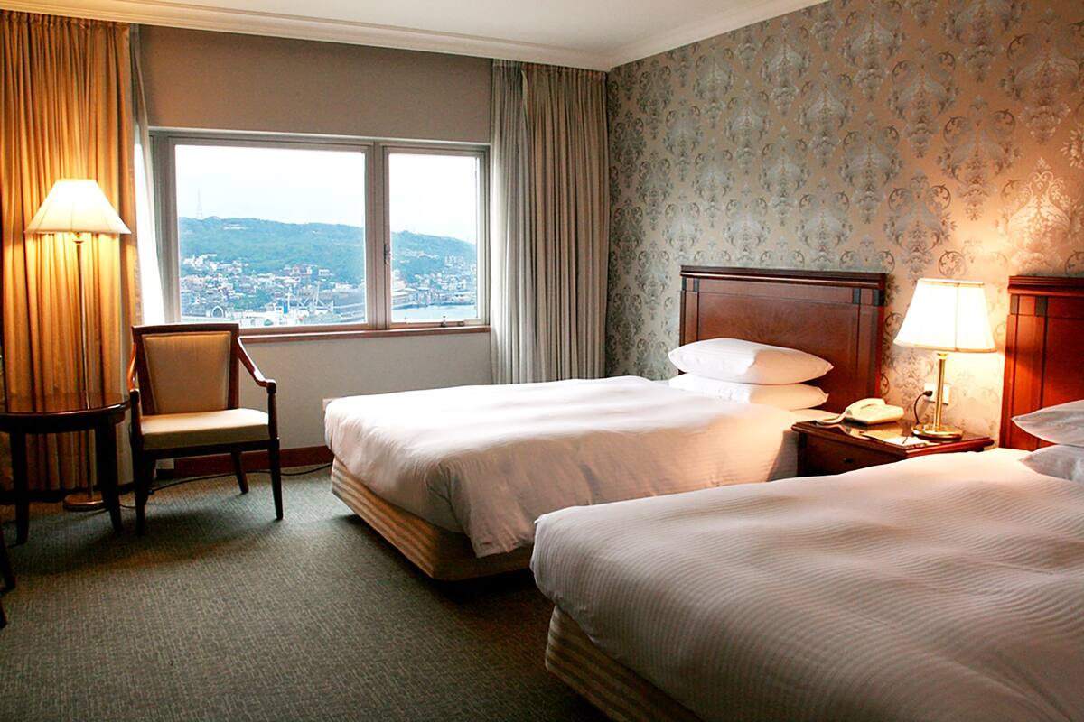 Hotelkamer met twee bedden, een stoel, een bureau met lamp en uitzicht op een stadsbeeld door een groot raam.