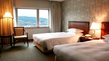 A hotel room with two beds, a chair, a desk with a lamp, and a view of a cityscape through a large window.