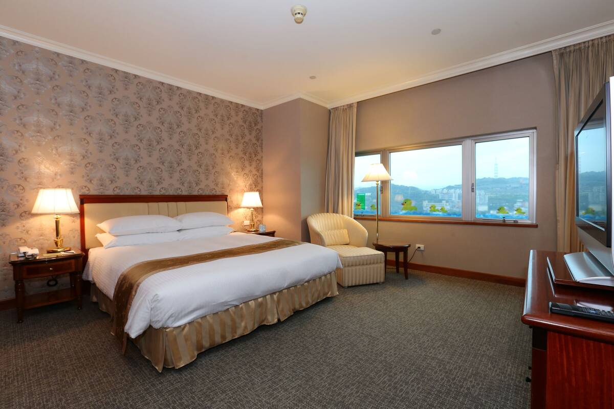 Hotelkamer met een groot bed, twee bedlampen, een stoel, een bureau met een televisie, en een raam met uitzicht op de stad.