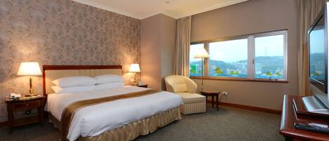 A hotel room with a large bed, two bedside lamps, a chair, a desk with a TV, and a window with a city view.