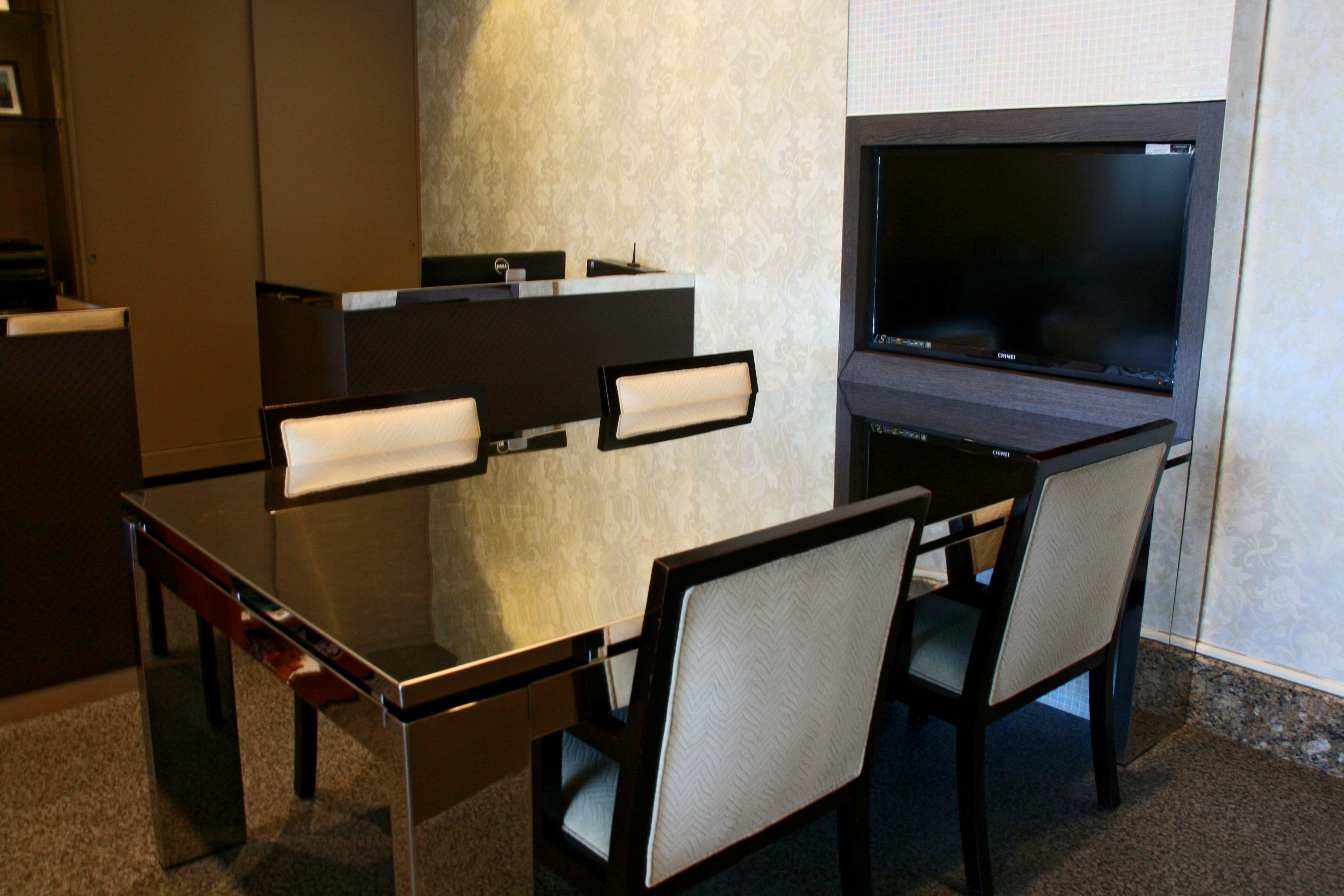 Une chambre d’hôtel moderne dotée d’un bureau en verre, de deux chaises, d’un téléviseur à écran plat et d’un petit espace bureau avec un ordinateur et un téléphone.