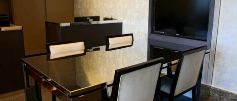 Une chambre d’hôtel moderne dotée d’un bureau en verre, de deux chaises, d’un téléviseur à écran plat et d’un petit espace bureau avec un ordinateur et un téléphone.