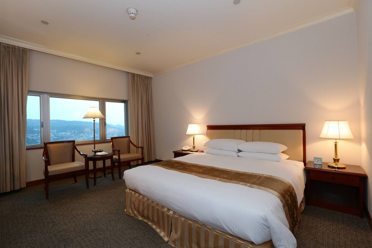 Een hotelkamer met een groot bed, twee nachtkastjes met lampen, een stoel, een kleine tafel en uitzicht op de stad door het raam.