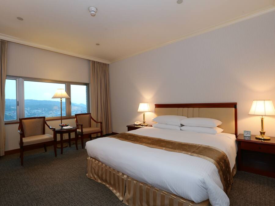 A hotel room with a large bed, two bedside tables with lamps, a chair, a small table, and a view of the city through the window.