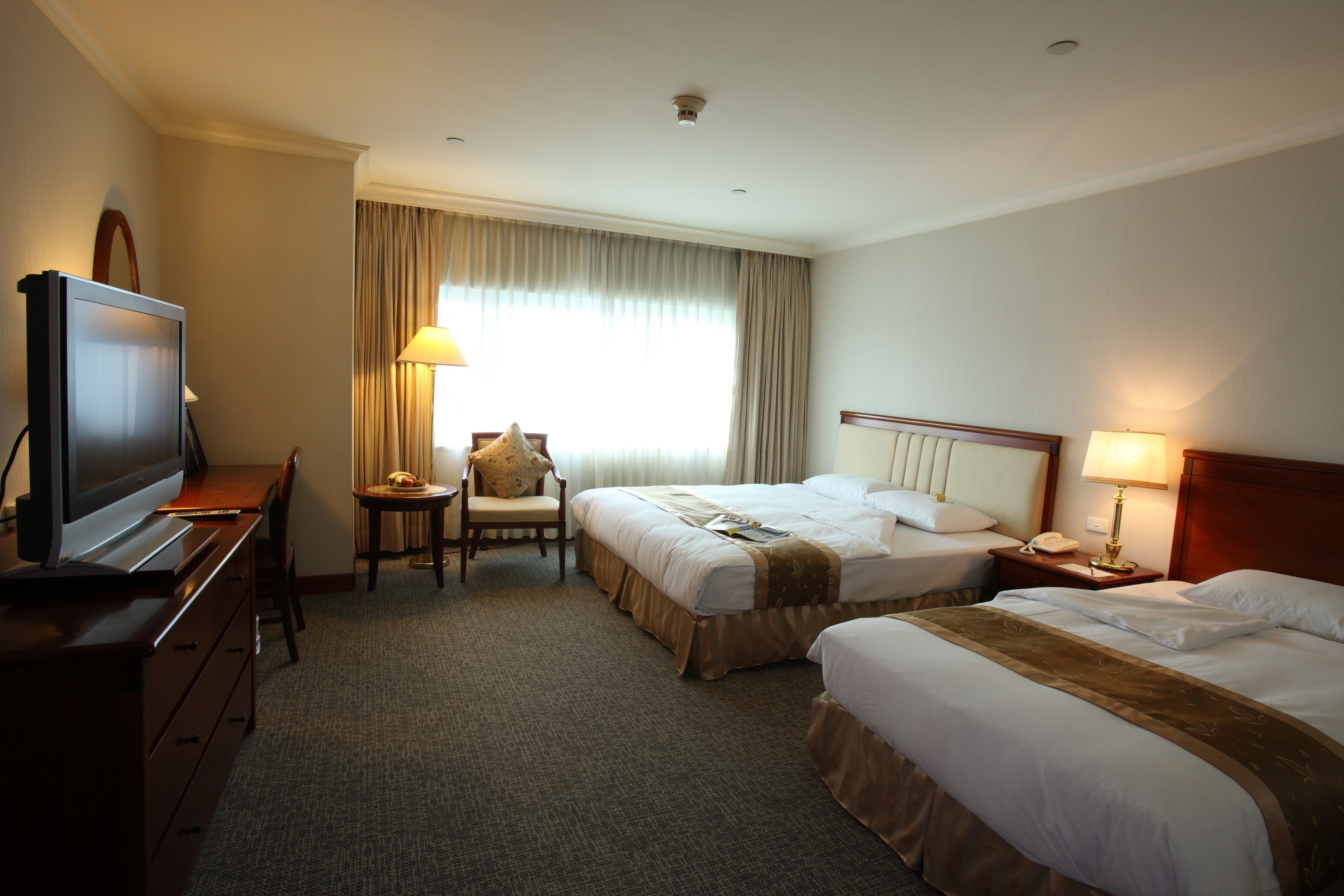 Habitación de hotel con dos camas, un escritorio con televisor, una silla, una mesita con lámpara y una ventana con cortinas.