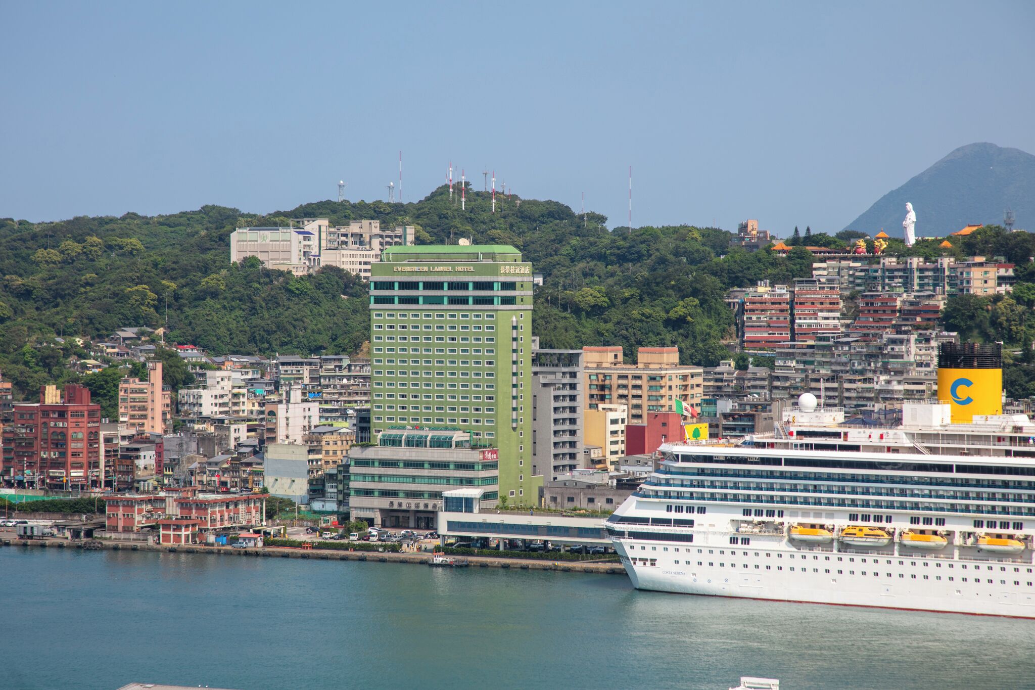 A cruise ship docked at a port with a cityscape in the background, including a prominent green building and a statue on a hill.