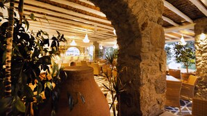 Interior com paredes de pedra, vigas de madeira, vasos de terracota, móveis de vime e luminárias pendentes.