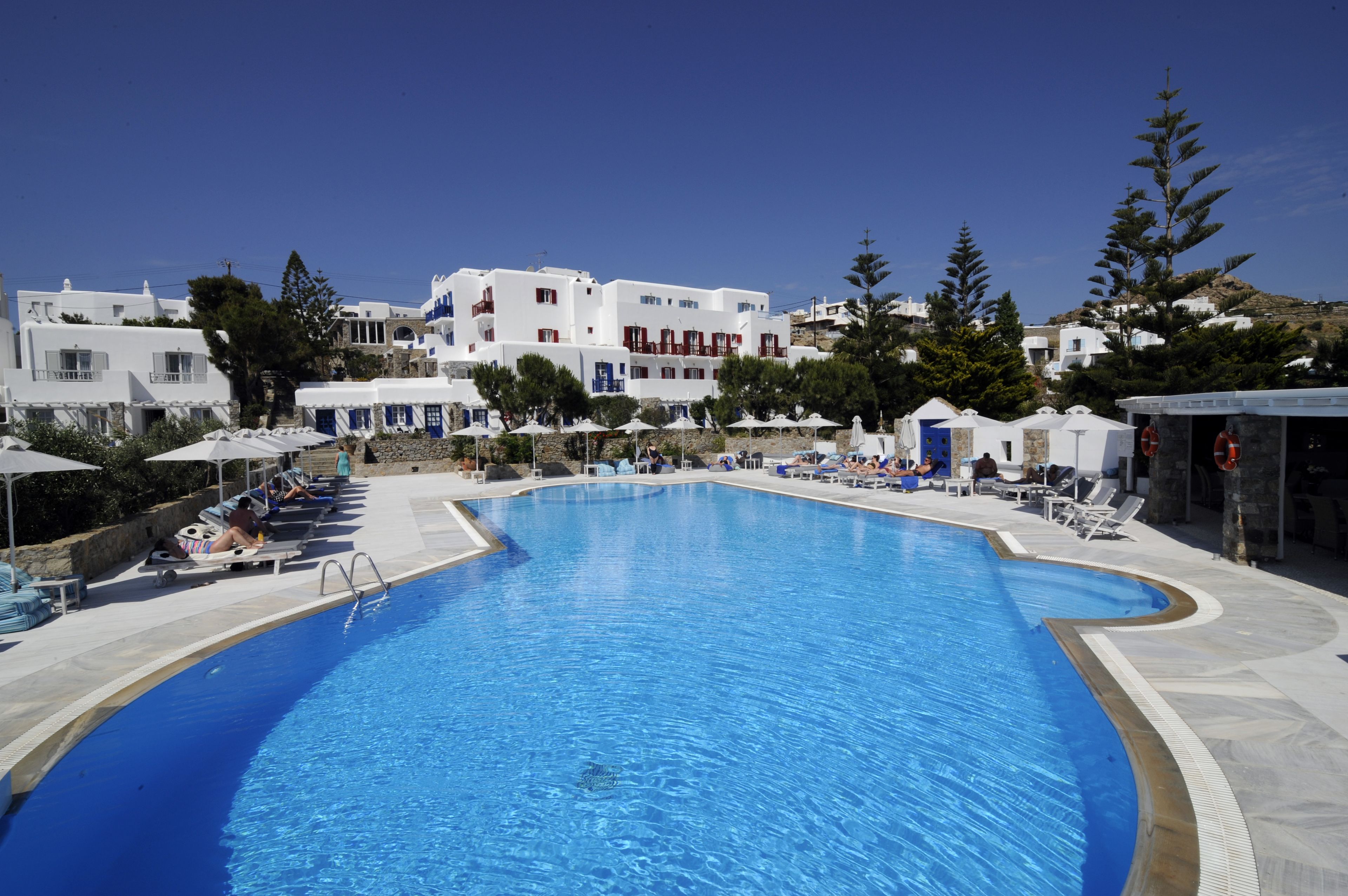 A large swimming pool with lounge chairs and umbrellas, surrounded by white buildings with red accents.