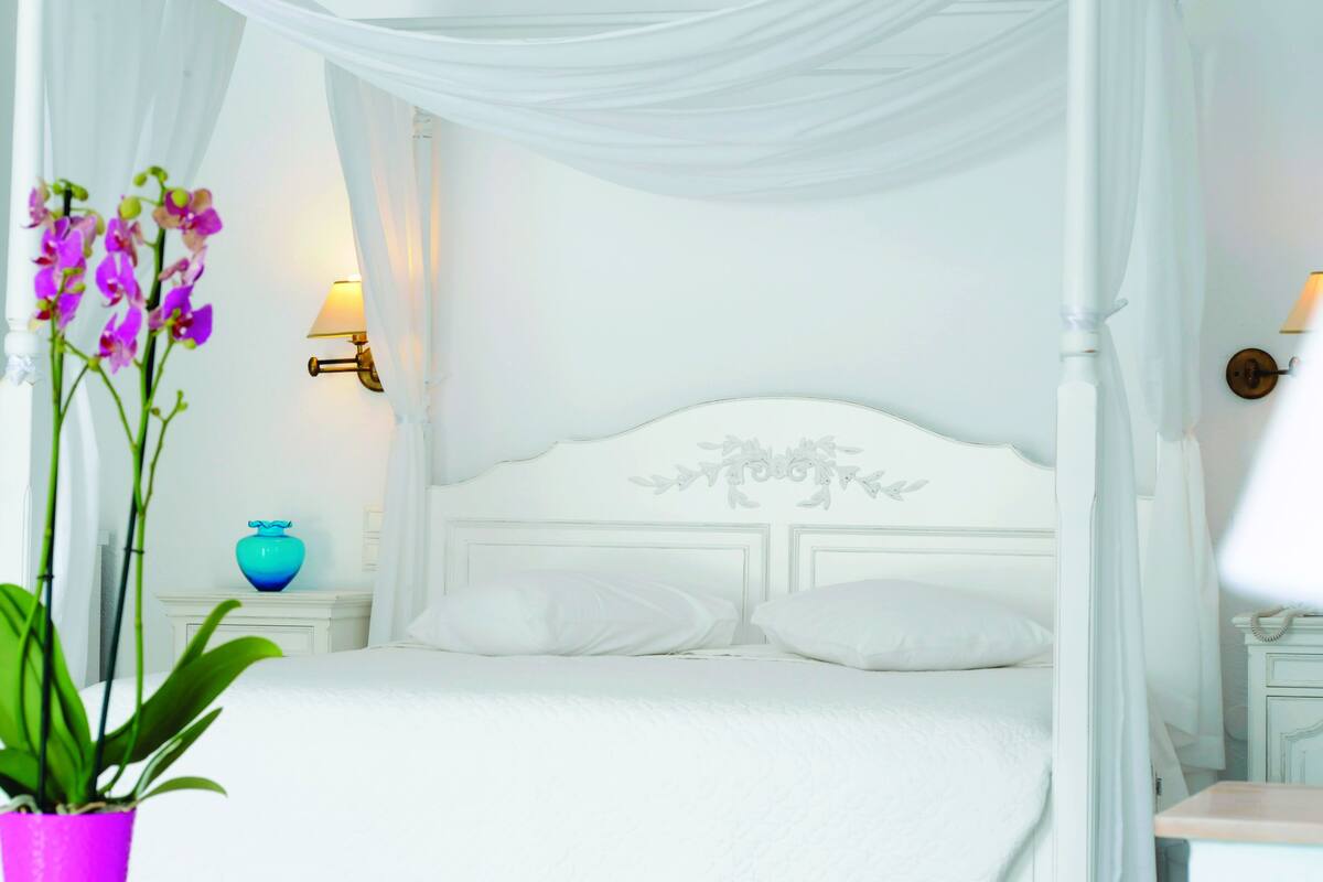 A white four-poster bed with a decorative headboard, white curtains, and a bedside table with a lamp.