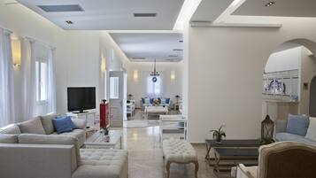 A spacious living room with a large sofa, a coffee table, and a television. There is a dining area with a table and chairs in the background.