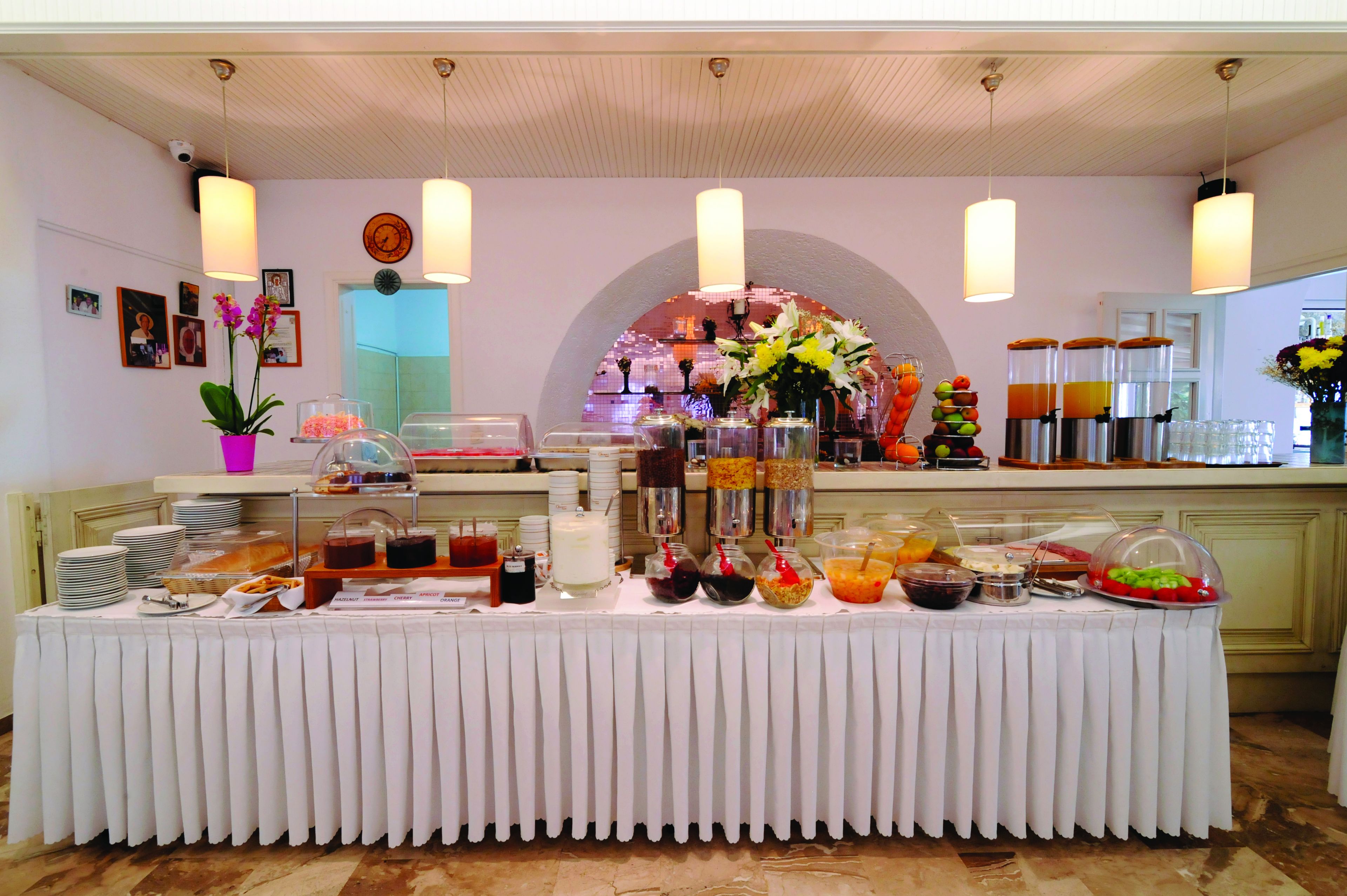 Disposición de buffet con diversos alimentos y bebidas, incluyendo frutas, ensaladas y refrescos, sobre una mesa con mantel blanco.