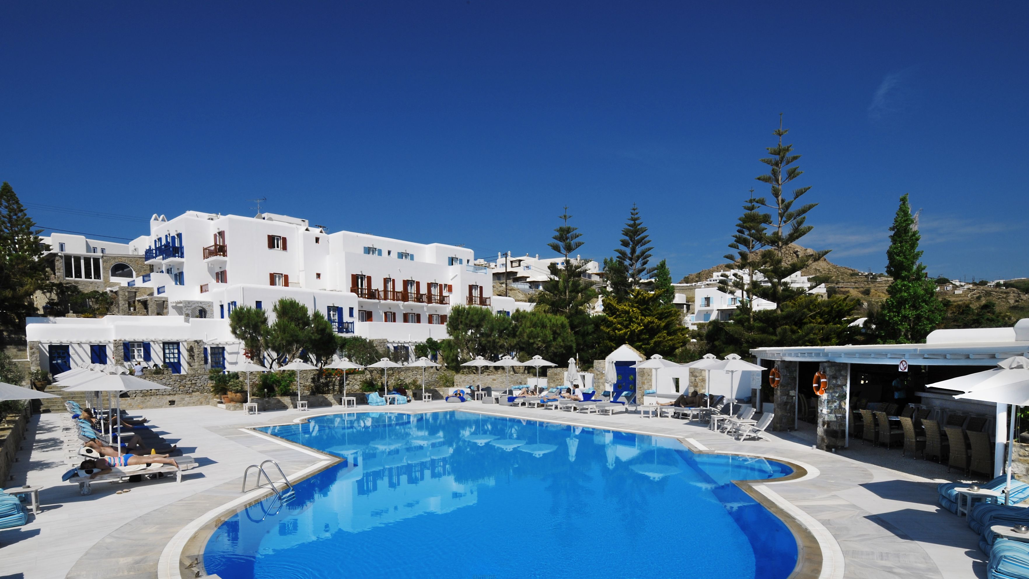 A pool area with lounge chairs and umbrellas, surrounded by white buildings with blue accents and trees.