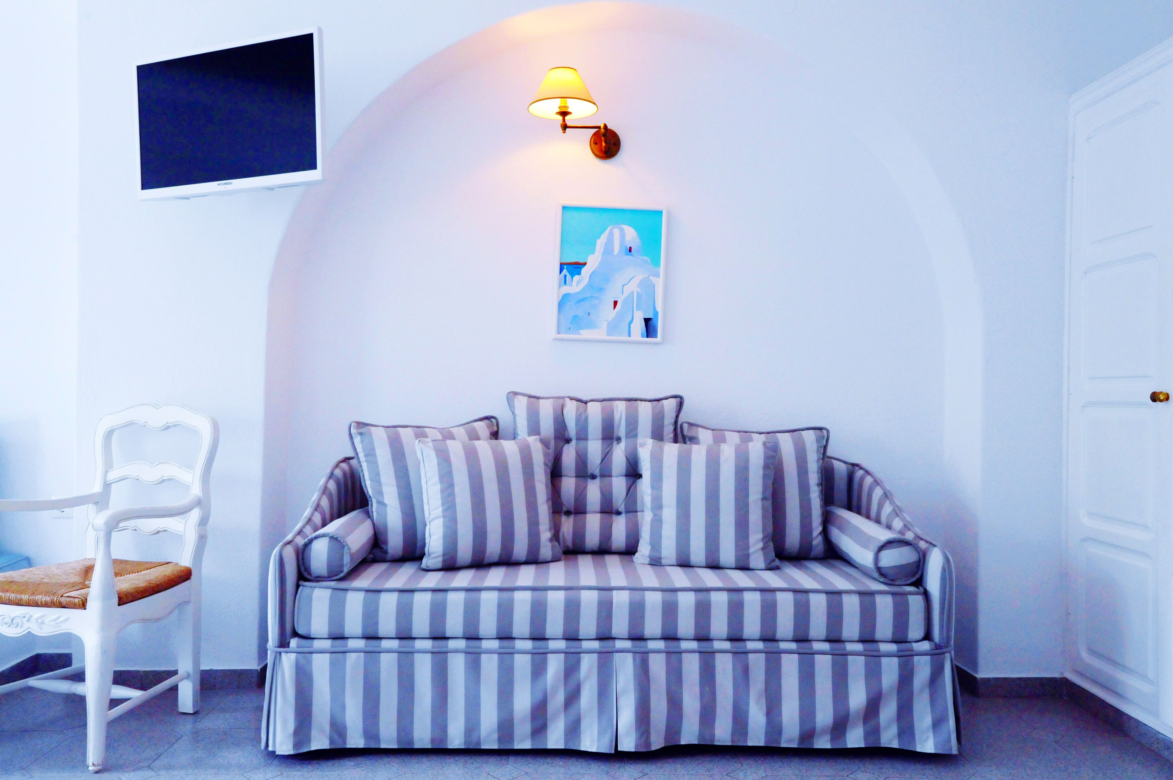 A white arched wall with a mounted flat-screen TV, a wall-mounted lamp, and a framed picture. A striped sofa with matching cushions is positioned against the wall, and a white chair with a brown cushion is to the left.