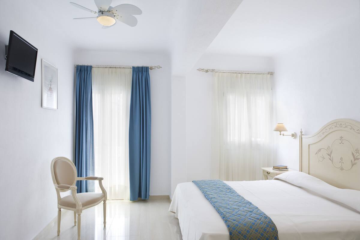 A hotel room with a bed, a chair, a bedside table with a lamp, and a ceiling fan.