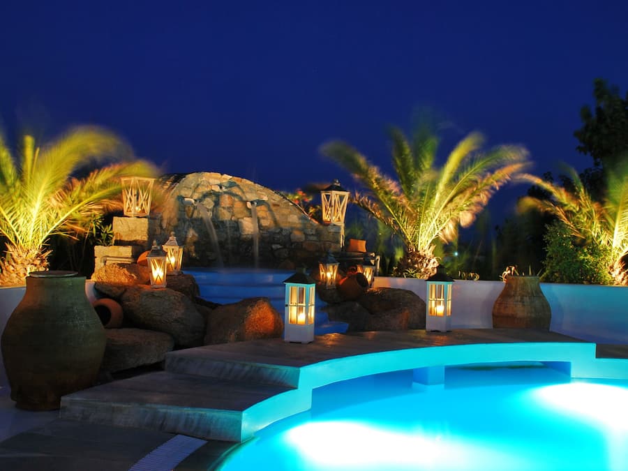 Un área de piscina nocturna con una estructura de piedra, faroles y palmeras.
