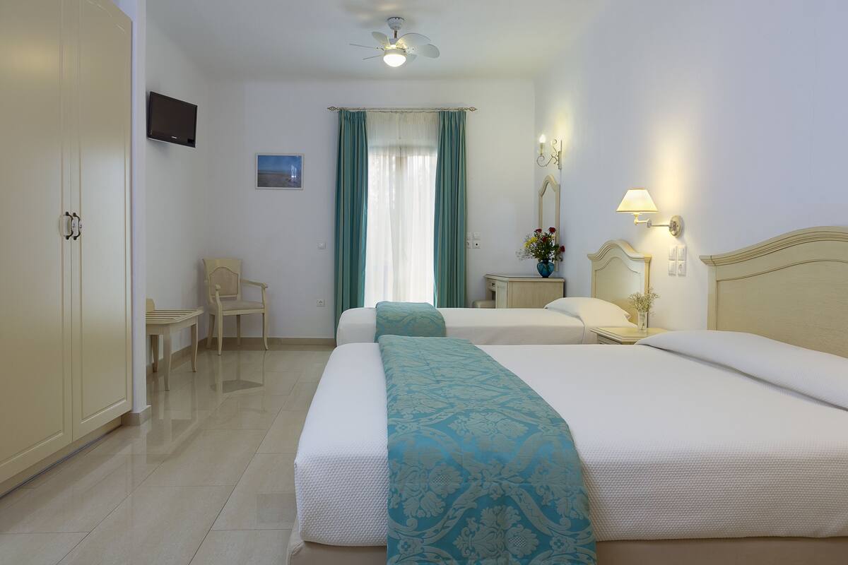 A hotel room with two beds, a television, a ceiling fan, a desk with a chair, and a window with curtains.