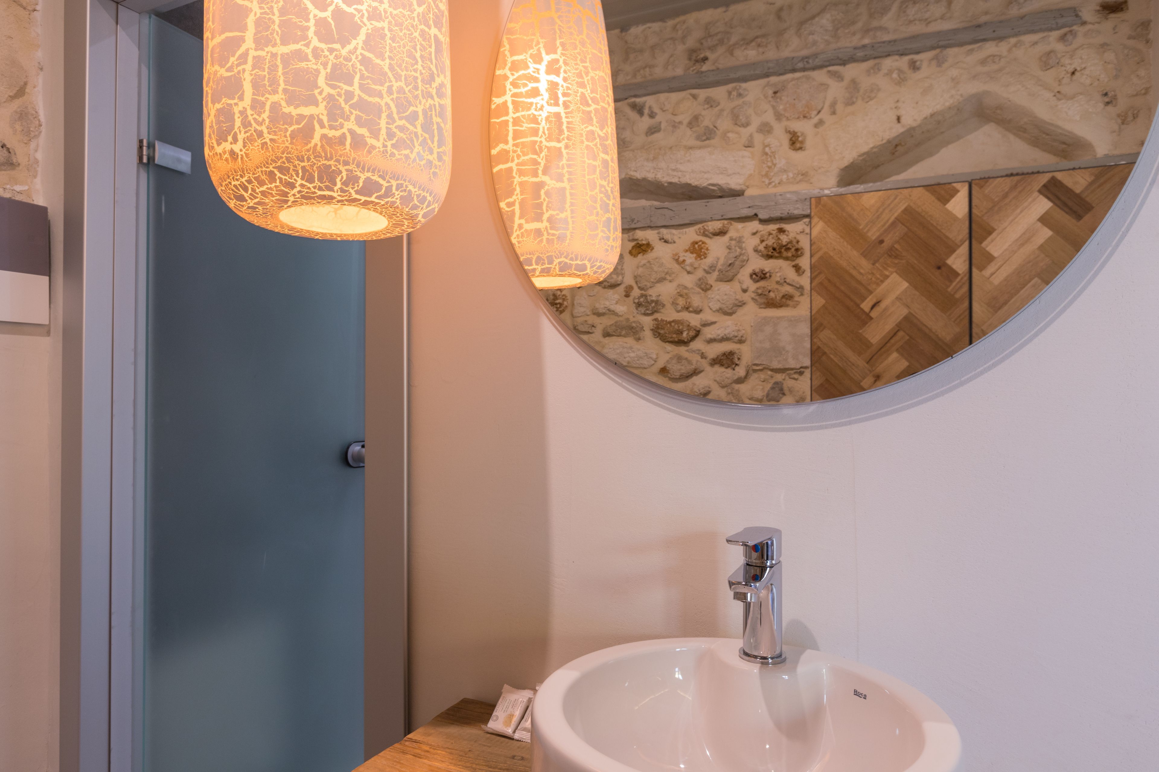 Een badkamer met een wit wastafelmeubel, een ronde spiegel en twee hanglampen met een gebarsten glazen design.