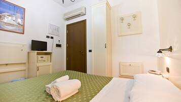 A hotel room with a green patterned bedspread, a neatly folded white towel, an air conditioning unit, a television, and a bedside table with a lamp.