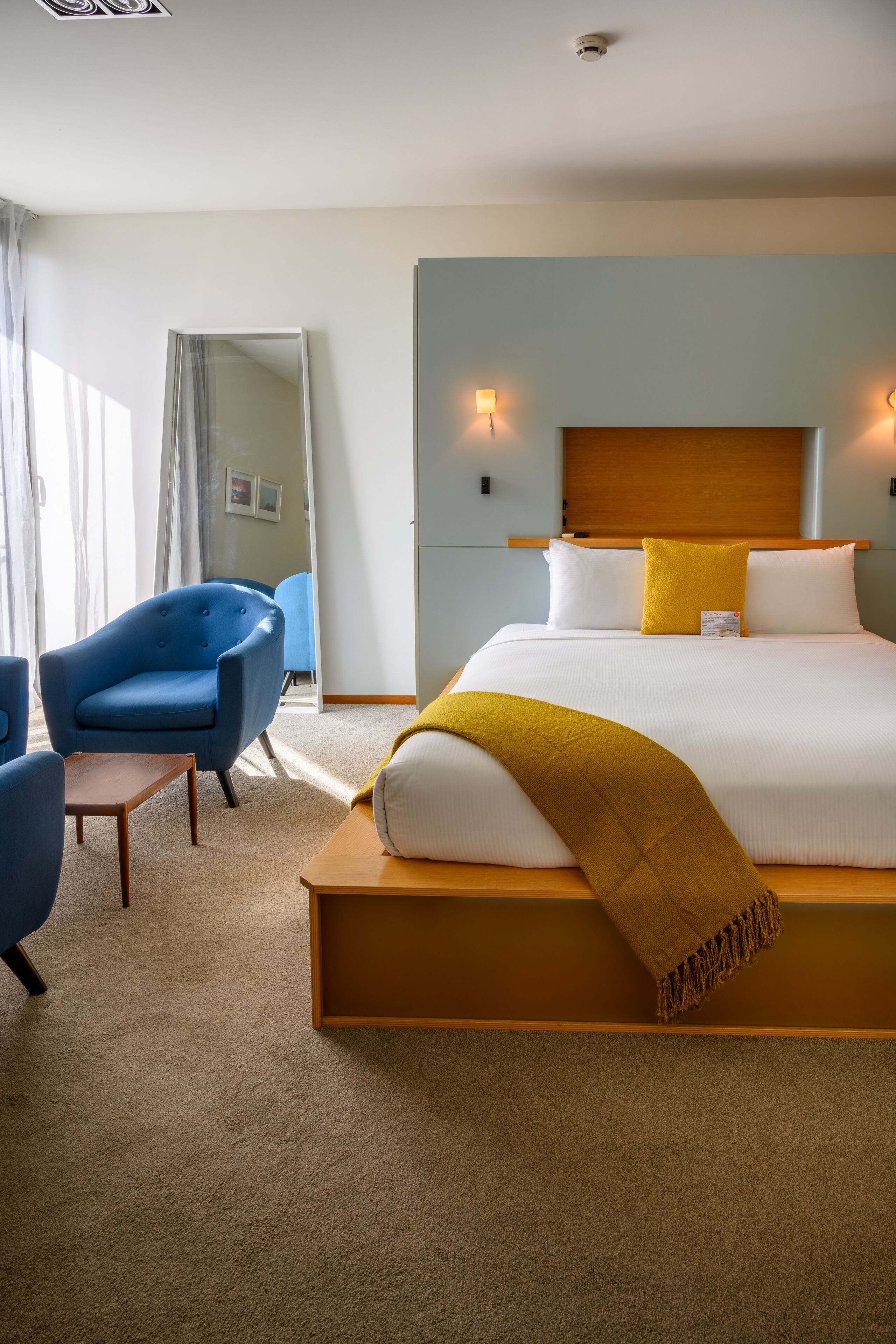Een hotelkamer met een bed, een blauwe fauteuil, een houten nachtkastje en een spiegel.