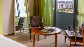 Quarto de hotel com cama, mesa redonda, duas cadeiras, espelho e vista da cidade através de amplas janelas.