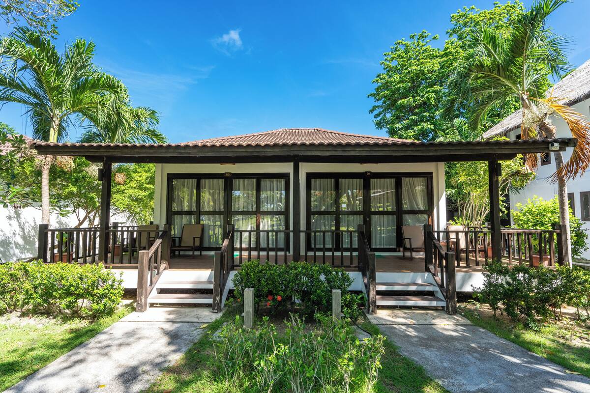 A covered outdoor seating area with a wooden deck, surrounded by lush greenery and palm trees.