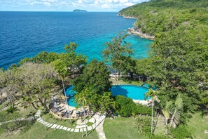 An aerial view of a resort with a swimming pool surrounded by lush greenery and a view of the ocean.
