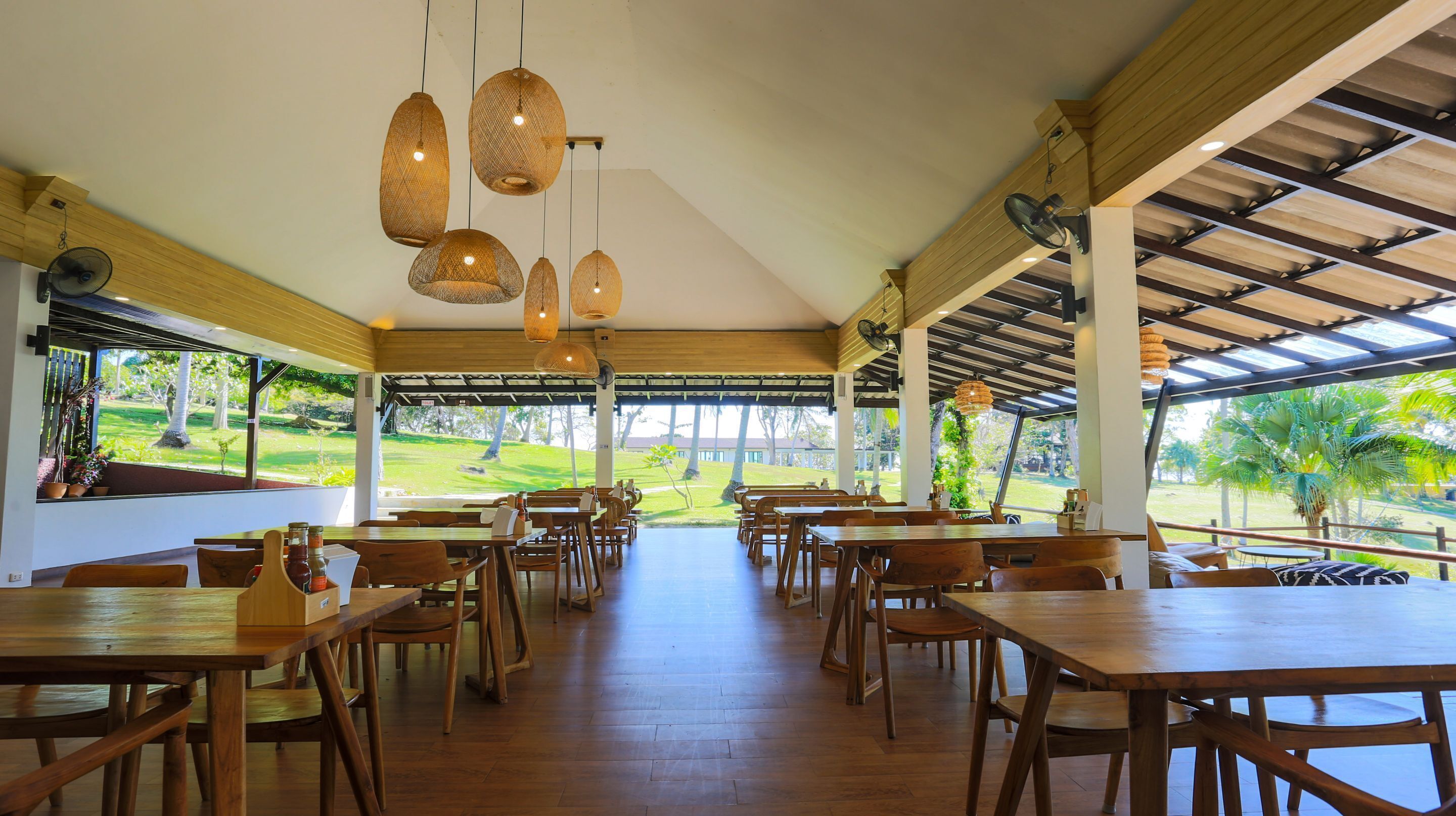 A spacious restaurant with wooden tables and chairs, hanging wicker light fixtures, and a view of a golf course through large windows.