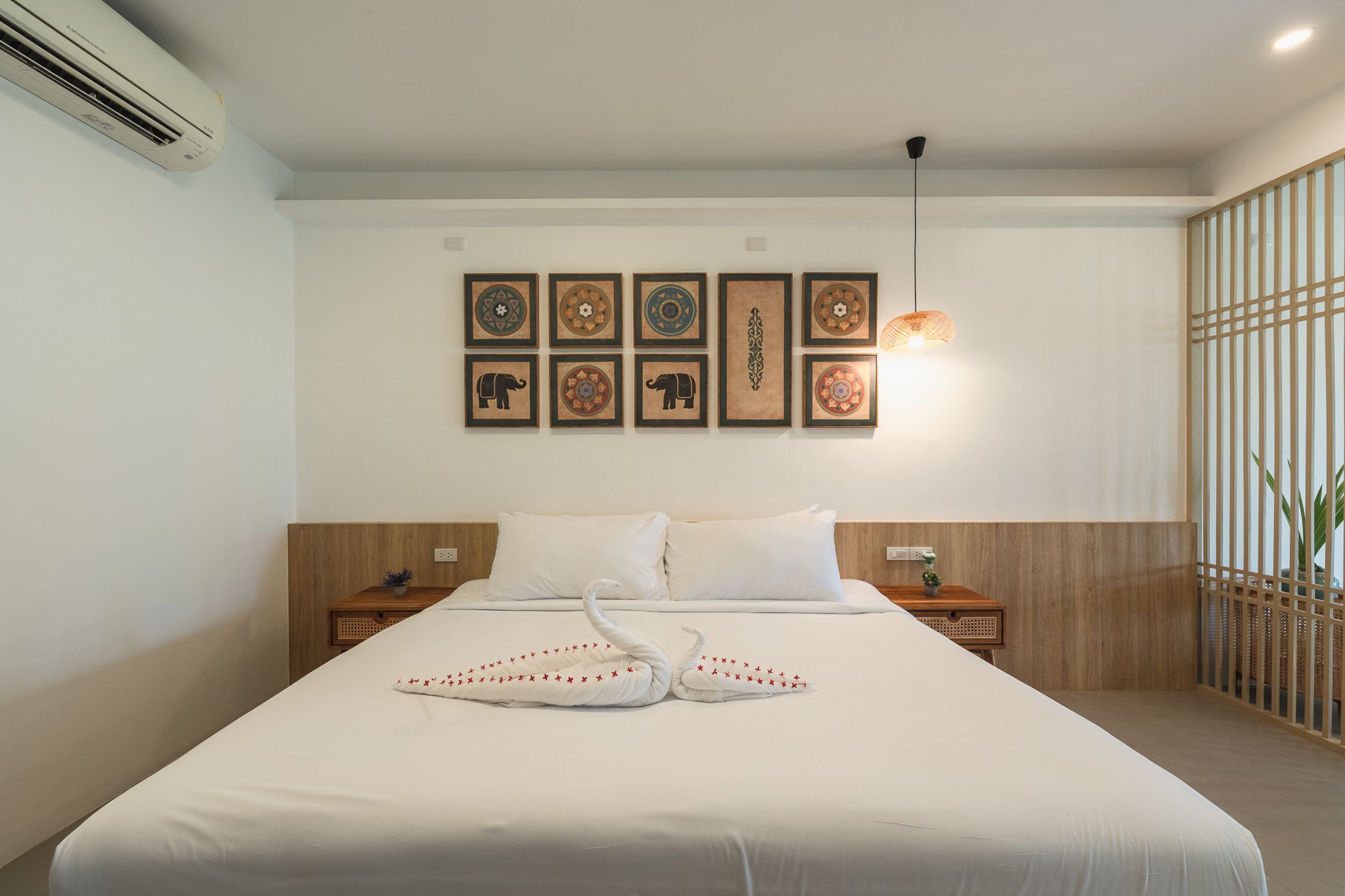 Habitación de hotel con una cama grande, aire acondicionado instalado en la pared, cabecero de madera, mesita de noche con lámpara y una pared decorativa con obras de arte enmarcadas.