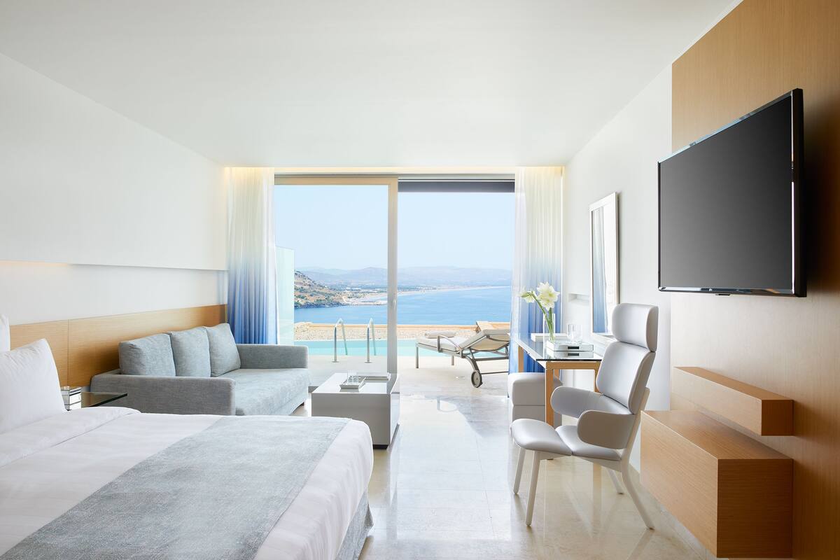 Habitación de hotel moderna con una cama grande, un sofá, un escritorio y un televisor. La habitación tiene vistas al mar y a las montañas.