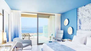 Una habitación de hotel moderna con una cama grande, una zona de estar con una silla y una mesa, vista al mar y un balcón con mesa y sillas.