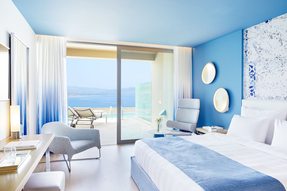 Una habitación de hotel moderna con una cama grande, una zona de estar con una silla y una mesa, vista al mar y un balcón con mesa y sillas.
