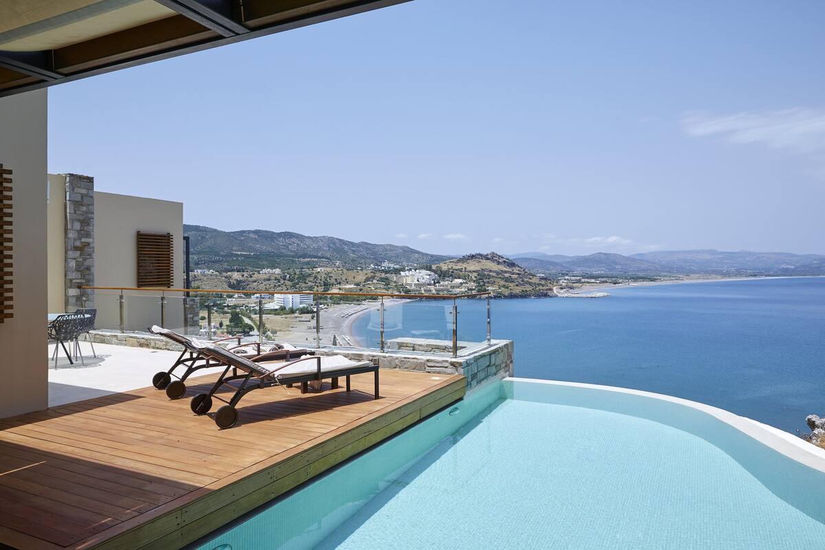 Zona junto a la piscina con terraza de madera, sillas de descanso y vistas al mar y a las montañas.