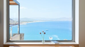 Habitación con amplia ventana que da a una playa y al mar. Se observa una bañera con vista al mar, un florero con flores y un vaso de agua en el alféizar.