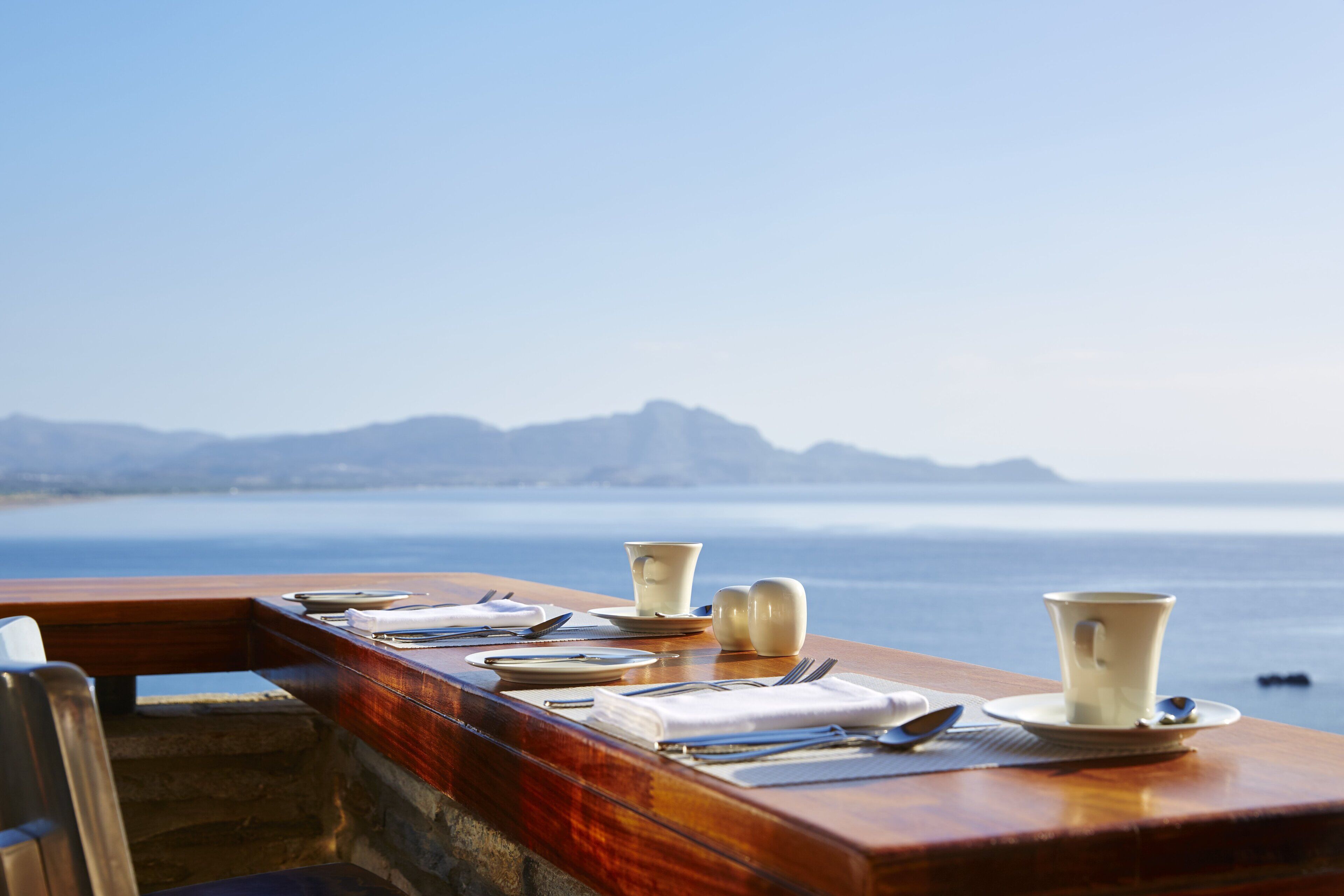 A table set with plates, cups, and cutlery overlooking a body of water with mountains in the distance.