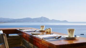 A table set with plates, cups, and cutlery overlooking a body of water with mountains in the distance.